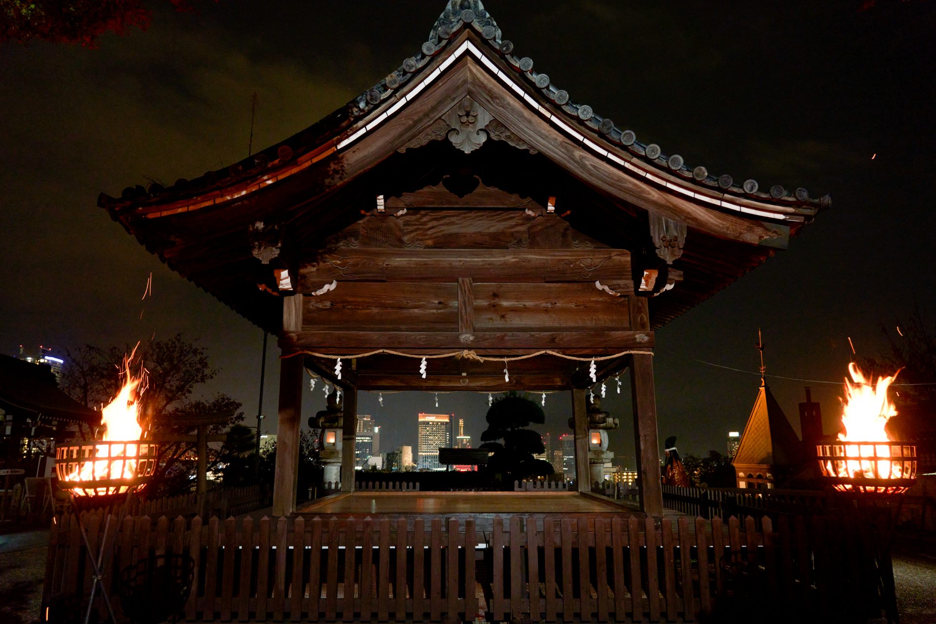 Un escenario de Noh iluminado por hogueras con la vista nocturna de Kobe al fondo